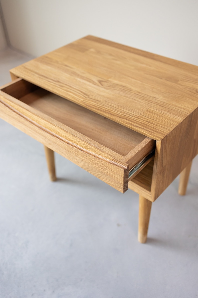 May include: A light wood nightstand with a single drawer that is open. The drawer has a simple design and is made of the same wood as the nightstand. The nightstand has four legs.