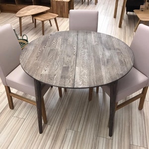 May include: A round wooden dining table with a gray finish and four beige chairs. The table has a leaf that can be added to extend the table.