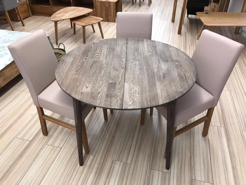 May include: A round wooden dining table with a gray finish and four beige chairs. The table has a leaf that can be added to extend the table.