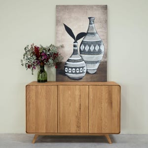 May include: A light wood cabinet with three doors. The cabinet has a rounded edge and sits on four legs. A painting of two vases with a geometric pattern hangs above the cabinet.