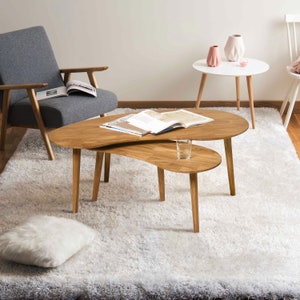 May include: A light wood coffee table with a unique, asymmetrical design. The table has two tiers, with the top tier being larger and the bottom tier being smaller. The table is sitting on a white shag rug.