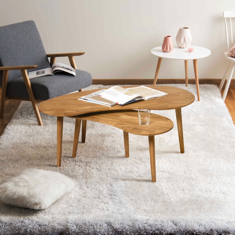 May include: A light wood coffee table with a unique, asymmetrical design. The table has two tiers, with the top tier being larger and the bottom tier being smaller. The table is sitting on a white shag rug.