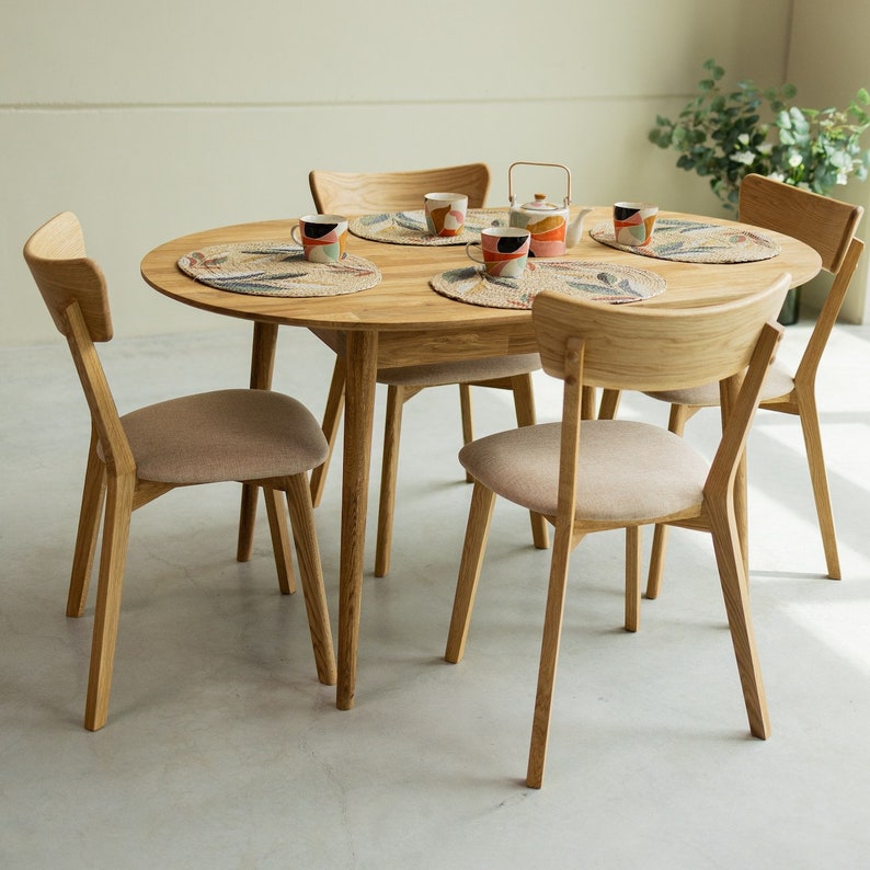 Pu&ograve; includere: Un tavolo da pranzo in legno con quattro sedie. Il tavolo ha un piano rotondo e una finitura in legno naturale. Le sedie sono anch'esse in legno e hanno una finitura marrone chiaro. Ci sono tovagliette all'americana sul tavolo con tazze colorate.