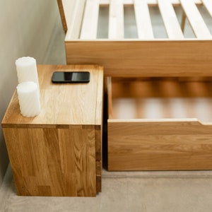 May include: A wooden nightstand with two drawers, a black cell phone, and two white candles. The nightstand is next to a wooden bed frame with a drawer.