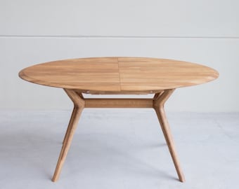 Puede incluir: Una mesa de madera ovalada de color marrón claro con una tapa dividida y cuatro patas. La mesa tiene un acabado de madera natural y está hecha de madera maciza.