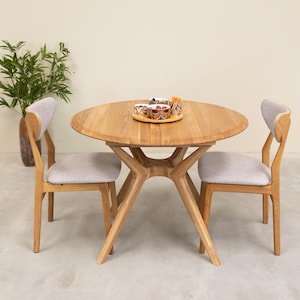 Può includere: Un tavolo da pranzo rotondo in legno chiaro con due sedie abbinate. Il tavolo ha una base dal design unico. Le sedie hanno sedute imbottite grigio chiaro e schienali curvi. Un vassoio con ciotole è sul tavolo.