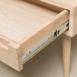 Puede incluir: Un caj&oacute;n de madera clara con un riel de caj&oacute;n de metal est&aacute; parcialmente abierto. El caj&oacute;n est&aacute; unido a una mesa de madera clara con una sola pata visible.