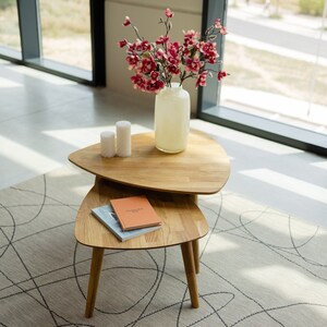 Puede incluir: Un conjunto de mesas nido de madera con dos tapas triangulares. La mesa est&aacute; hecha de madera marr&oacute;n claro y tiene un acabado natural. La mesa est&aacute; decorada con un jarr&oacute;n de flores rosas y dos velas blancas.