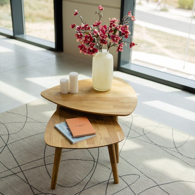Puede incluir: Un conjunto de mesas nido de madera con dos tapas triangulares. La mesa est&aacute; hecha de madera marr&oacute;n claro y tiene un acabado natural. La mesa est&aacute; decorada con un jarr&oacute;n de flores rosas y dos velas blancas.