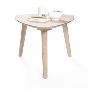Puede incluir: Una mesa de centro triangular de madera clara con tres patas. La mesa tiene un taz&oacute;n blanco con art&iacute;culos decorativos y un libro en la parte superior.