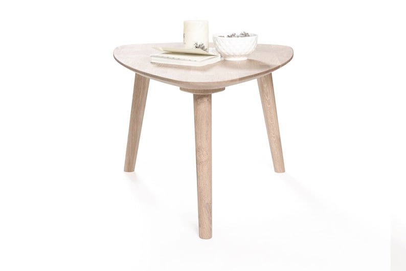 Puede incluir: Una mesa de centro triangular de madera clara con tres patas. La mesa tiene un taz&oacute;n blanco con art&iacute;culos decorativos y un libro en la parte superior.