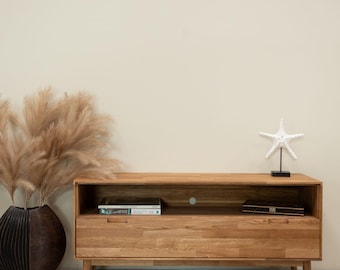 Könnte beinhalten: Ein heller Holz-TV-Schrank mit einer Schublade und einem offenen Regal. Der Schrank hat eine weiße Seestern-Dekoration oben und ein Buch im Regal. Der Schrank steht vor einer hellbeigen Wand.