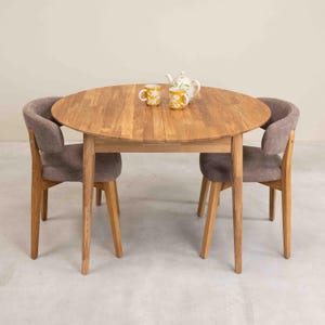 Op de afbeelding: Een ronde houten tafel met twee stoelen. De tafel heeft een lichte houtkleur en de stoelen hebben grijze stoffen zittingen. Er staan twee gele mokken en een witte theepot op de tafel.