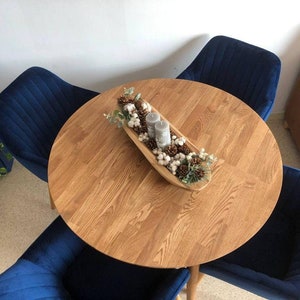 May include: A round wooden table with a natural wood finish. The table is set with a white bowl filled with cotton balls, pine cones, and candles. Four blue velvet chairs surround the table.