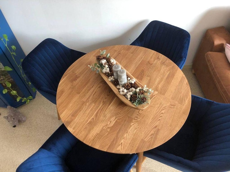 May include: A round wooden table with a natural wood finish. The table is set with a white bowl filled with cotton balls, pine cones, and candles. Four blue velvet chairs surround the table.