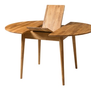 May include: A round wooden table with a leaf extension. The table has a light brown finish and four legs. The leaf is open and propped up against the table.