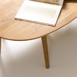 May include: A light wood oval coffee table with four slender legs. The table has a natural wood finish and is topped with a magazine.