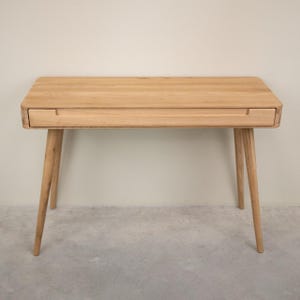 Peut inclure: Un bureau en bois clair avec un seul tiroir et quatre pieds inclinés. Le bureau a un dessus rectangulaire avec des coins arrondis. Le tiroir a une poignée encastrée. Le bureau est en bois de couleur claire.