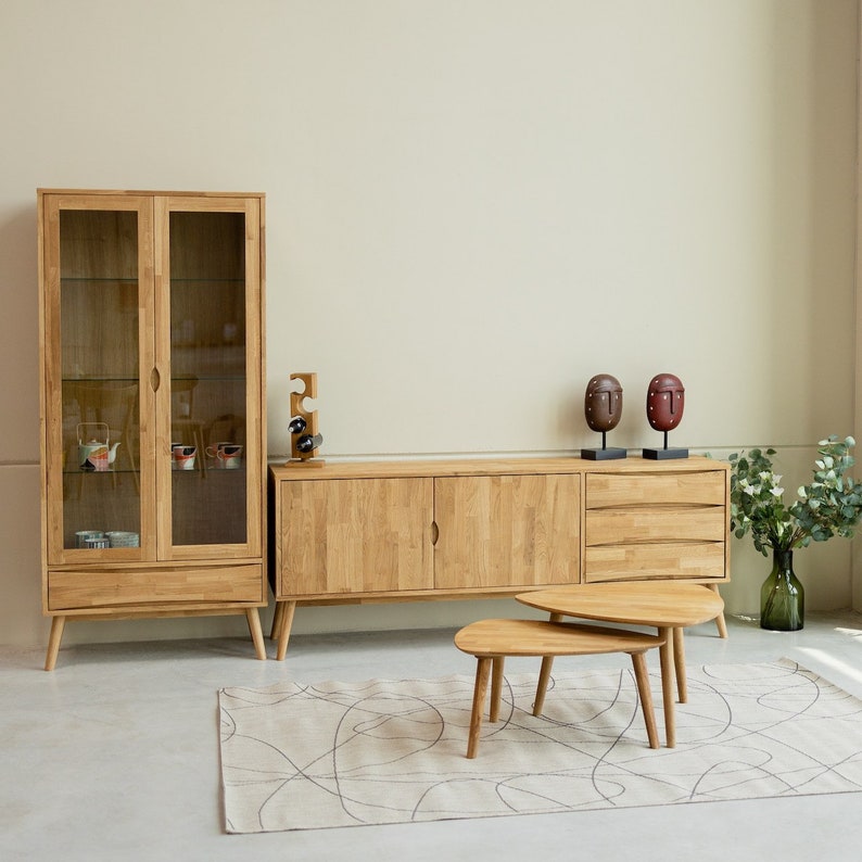 Puede incluir: Un armario de madera clara con puertas de vidrio y cajones, un aparador de madera clara a juego con cajones y dos mesas nido con acabado en madera clara. El armario y el aparador est&aacute;n contra una pared blanca, y las mesas est&aacute;n sobre una alfombra con un dise&ntilde;o abstracto marr&oacute;n claro y blanco.