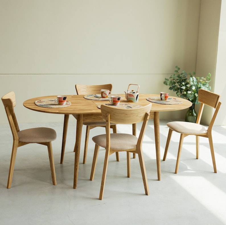 Pu&ograve; includere: Un tavolo da pranzo ovale in legno chiaro con quattro sedie. Il tavolo ha una finitura in legno naturale e le sedie hanno un sedile in tessuto marrone chiaro.