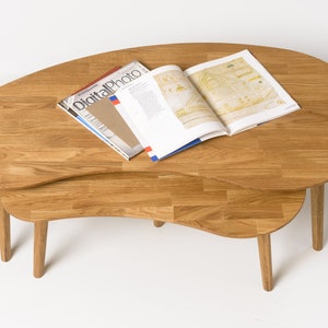 May include: A set of two wooden nesting coffee tables with a unique, organic shape. The tables are made of light brown wood and have a natural finish. The top table is slightly larger than the bottom table and has a rounded edge. The tables are shown with a magazine on top.