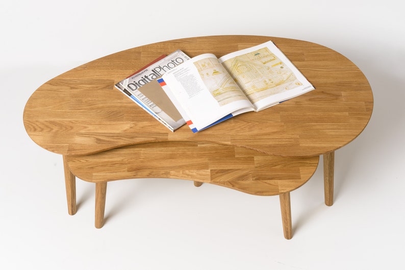 May include: A set of two wooden nesting coffee tables with a unique, organic shape. The tables are made of light brown wood and have a natural finish. The top table is slightly larger than the bottom table and has a rounded edge. The tables are shown with a magazine on top.