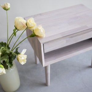 May include: A white wooden nightstand with a drawer and a shelf. The nightstand has a light wood grain and is painted white. The nightstand is sitting on a light gray surface.