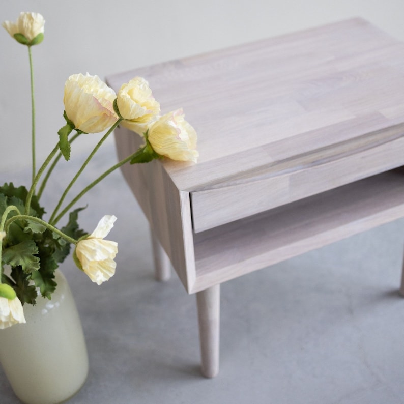 May include: A white wooden nightstand with a drawer and a shelf. The nightstand has a light wood grain and is painted white. The nightstand is sitting on a light gray surface.