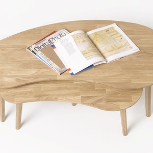 May include: A set of two wooden nesting coffee tables with a unique, organic shape. The tables are made of light brown wood and have a natural finish. The top table has a magazine on it.