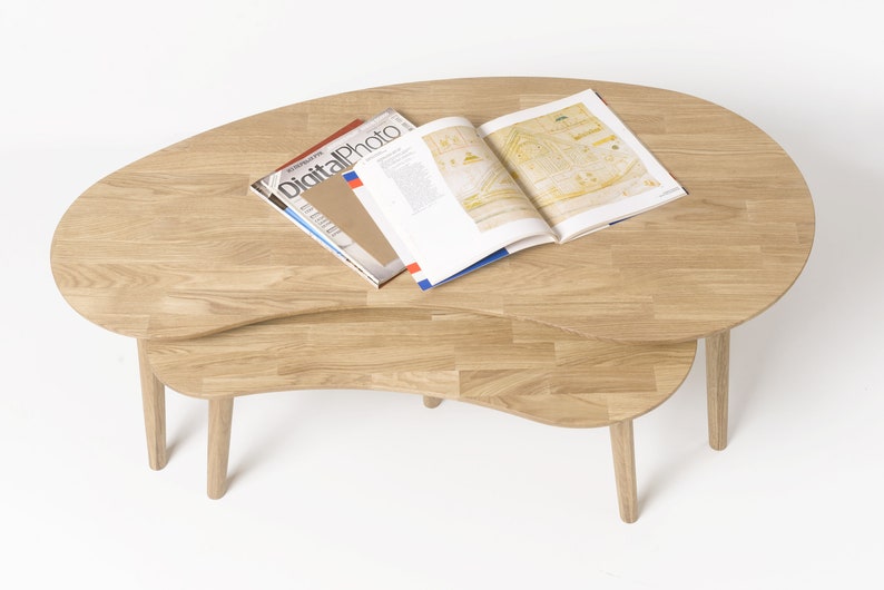 May include: A set of two wooden nesting coffee tables with a unique, organic shape. The tables are made of light brown wood and have a natural finish. The top table has a magazine on it.