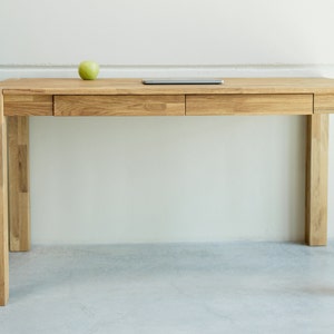Può includere: Una scrivania in legno chiaro con due cassetti e un design semplice. La scrivania ha un piano rettangolare e quattro gambe.