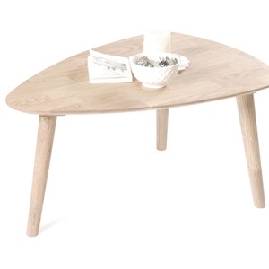 Puede incluir: Una mesa de centro triangular de madera clara con tres patas. La mesa tiene un taz&oacute;n blanco con art&iacute;culos decorativos en la parte superior.