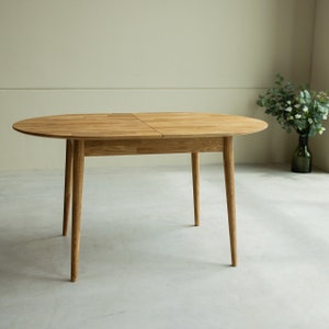 Pu&ograve; includere: Un tavolo da pranzo ovale in legno chiaro con un'estensione a foglia. Il tavolo ha quattro gambe e una finitura in legno naturale.