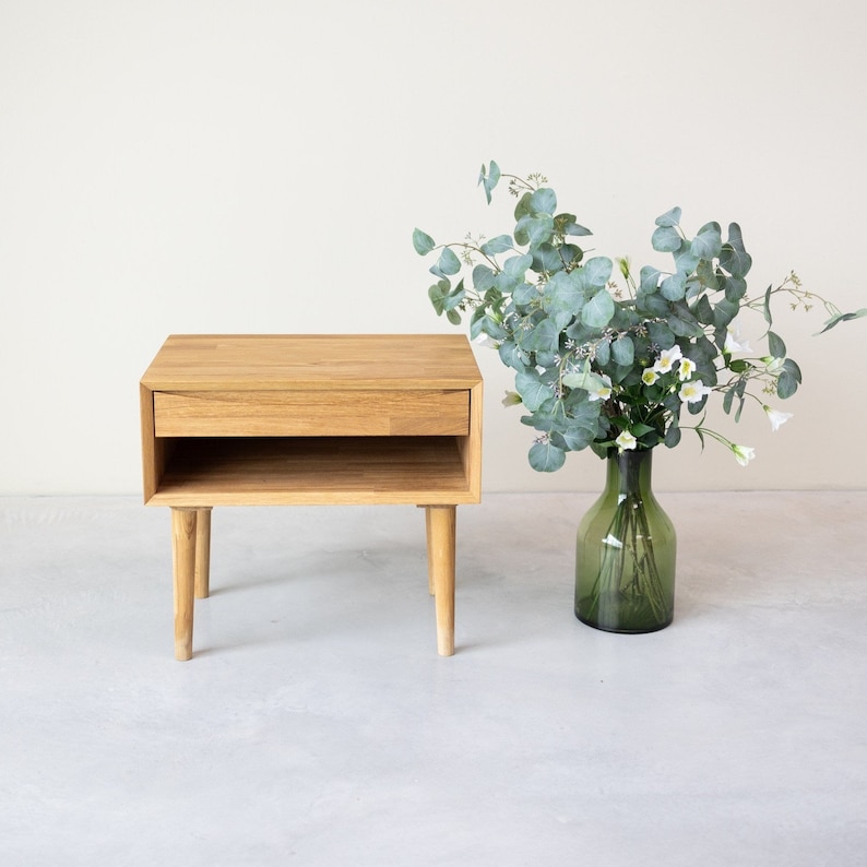May include: A light wood nightstand with a drawer and a shelf. The nightstand has four legs and is sitting on a white surface. A vase with green leaves and white flowers is next to the nightstand.
