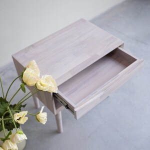 May include: White wooden nightstand with a drawer that is open. The nightstand has a simple design and is made of solid wood. The drawer is made of the same wood and has a smooth finish.