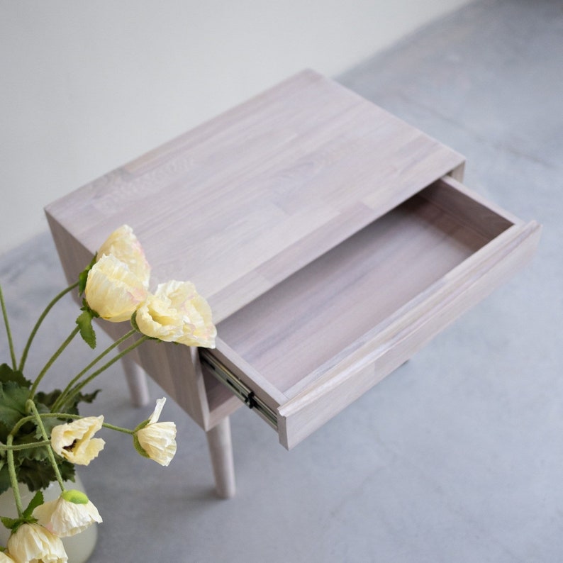May include: White wooden nightstand with a drawer that is open. The nightstand has a simple design and is made of solid wood. The drawer is made of the same wood and has a smooth finish.