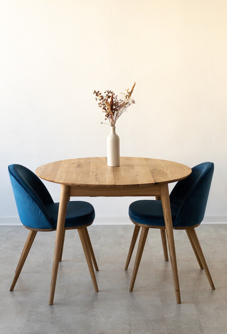 May include: A round wooden dining table with two blue velvet chairs. The table is set with a white vase filled with dried flowers.