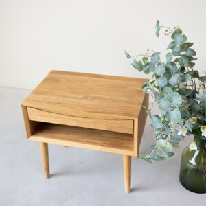 May include: A light wood nightstand with a single drawer and a lower shelf. The nightstand has tapered legs and a curved drawer front.