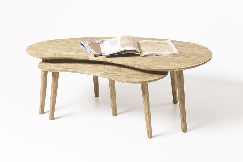 May include: Two wooden nesting coffee tables with a unique, organic shape. The top table has a magazine and a book on it.