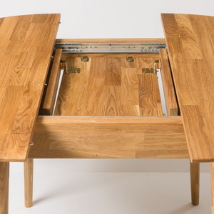 May include: A round wooden dining table with two leaves that extend out from the center. The table is made of light brown wood and has a natural finish. The leaves are supported by metal tracks and hinges.