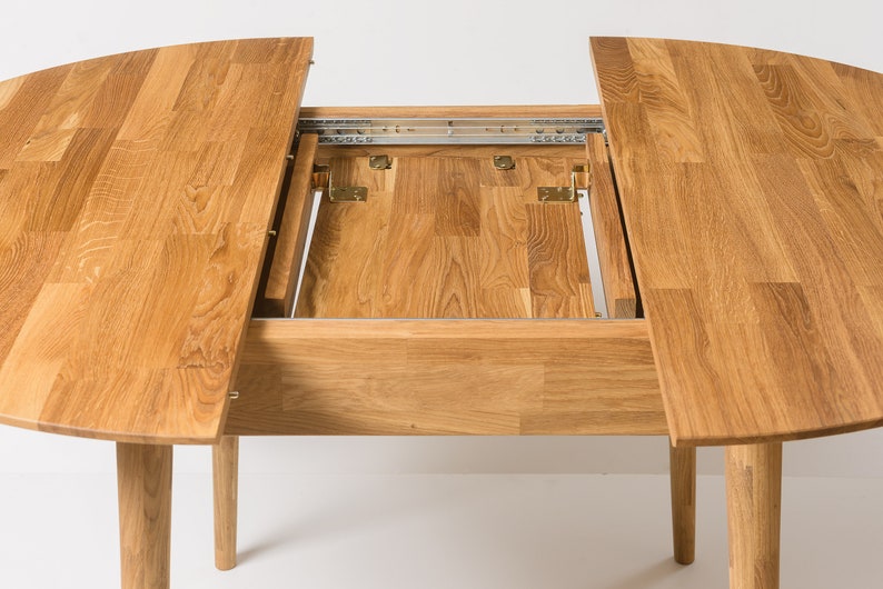 May include: A round wooden dining table with two leaves that extend out from the center. The table is made of light brown wood and has a natural finish. The leaves are supported by metal tracks and hinges.