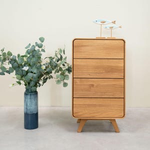 Puede incluir: Una cómoda de madera clara con cuatro cajones y bordes redondeados. La cómoda está sobre un suelo gris claro y tiene un par de peces decorativos de madera en la parte superior.