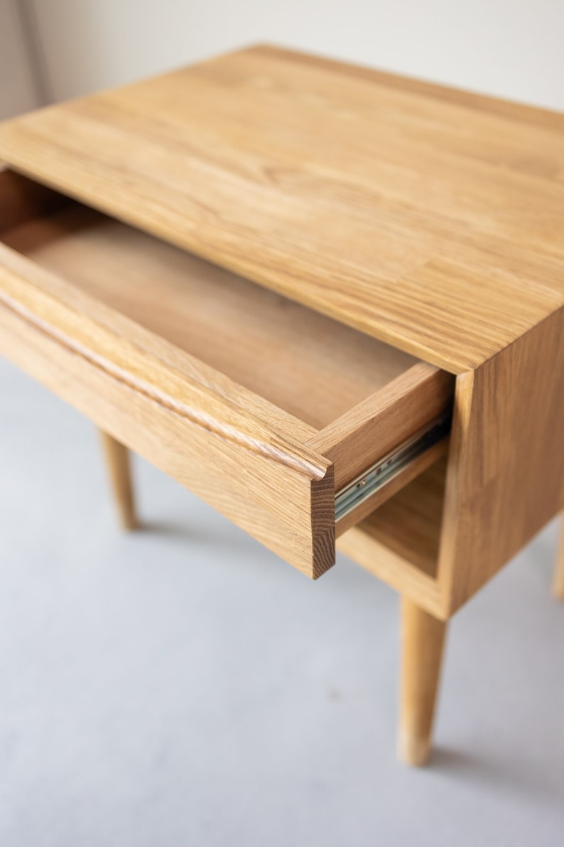 May include: A light wood nightstand with a single drawer that is open, revealing the interior. The drawer is made of the same light wood and has a smooth finish. The nightstand has four legs that are tapered and slightly angled.