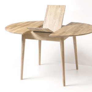 May include: A round wooden table with a leaf extension that can be pulled out to expand the table's size. The table has a light wood finish and four slender legs.
