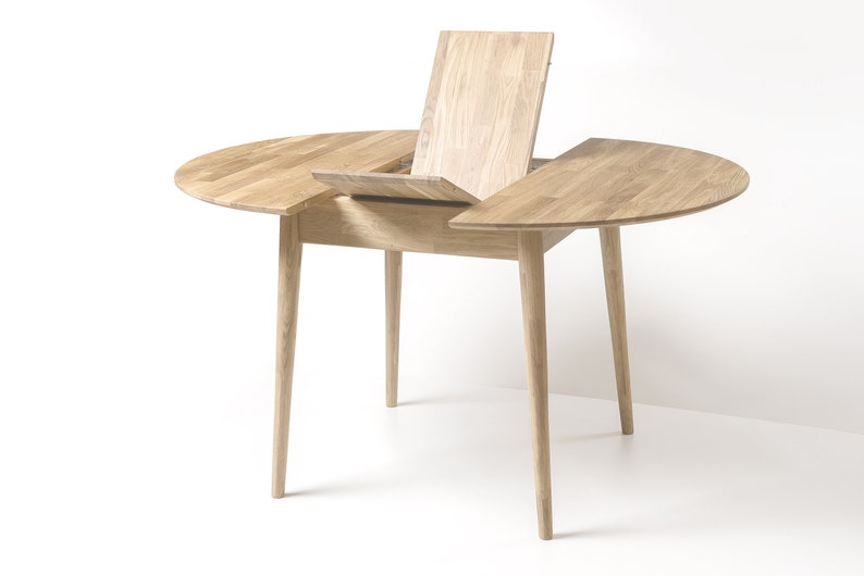 May include: A round wooden table with a leaf extension that can be pulled out to expand the table's size. The table has a light wood finish and four slender legs.