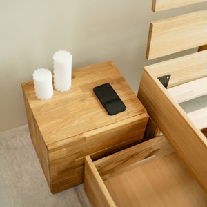 May include: A wooden nightstand with two drawers and a top surface. The nightstand has a natural wood finish and is made of solid wood. There are two white candles and a black smartphone on the top surface.