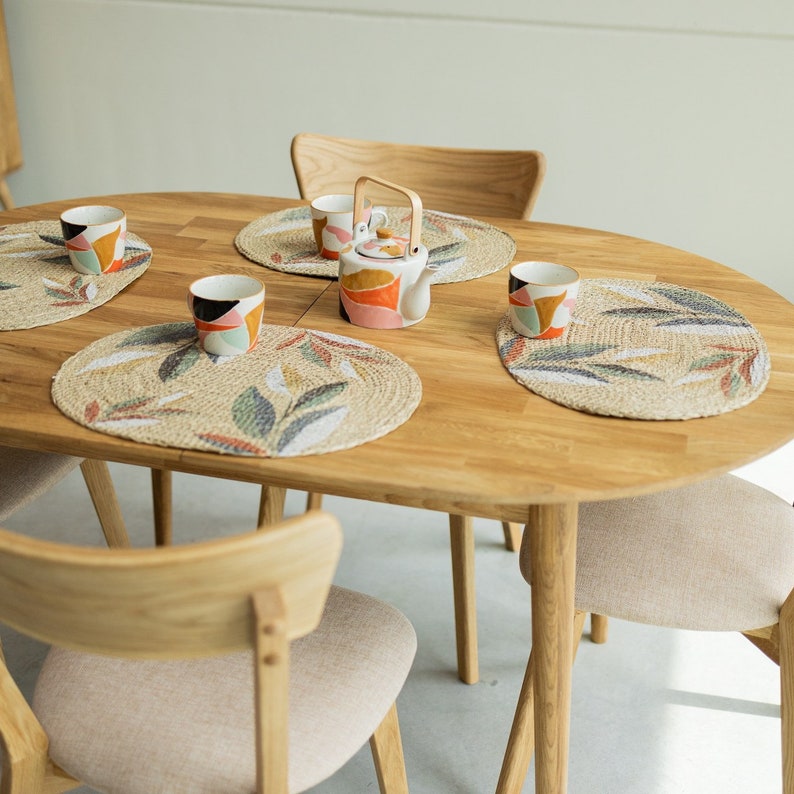 Pu&ograve; includere: Un tavolo ovale in legno con quattro sedie, apparecchiato per quattro persone con tazze colorate e una teiera su sottobicchieri intrecciati. Il tavolo ha una finitura in legno naturale e le sedie hanno telai in legno chiaro con cuscini beige.
