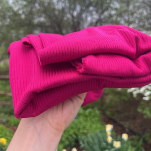 May include: A folded, hot pink ribbed knit fabric. The fabric is held in a hand, with the edges of the fabric visible. The background is a blurred outdoor scene with greenery and a wooden fence.