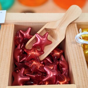 May include: A wooden box containing numerous shiny, red star-shaped items. A small wooden scoop sits amongst the stars. The stars have a glossy finish and appear translucent.