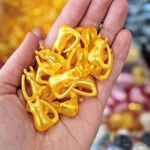 K&ouml;nnte beinhalten: Eine Handvoll kleiner, goldener Kapseln, wahrscheinlich mit einem Sch&ouml;nheitsserum oder einem Nahrungserg&auml;nzungsmittel gef&uuml;llt. Die Kapseln haben die Form kleiner, abgerundeter Figuren.
